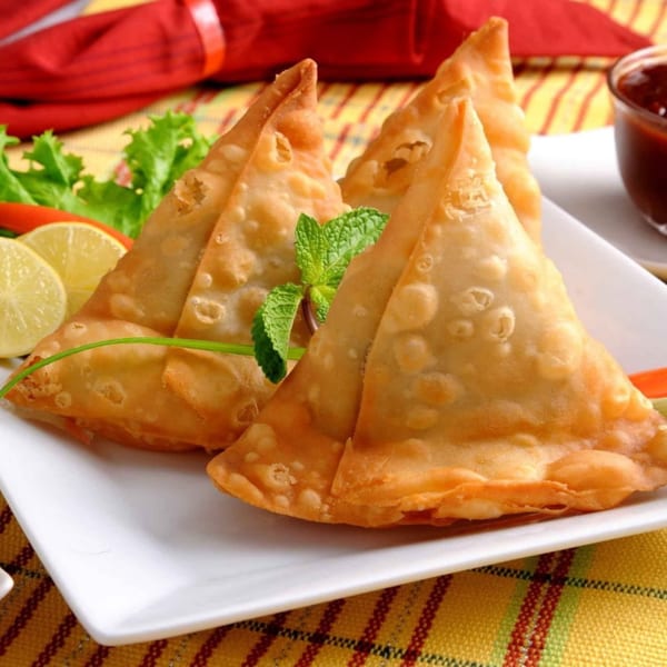 Golden-brown Bawarchi Biryanis samosas garnished with mint, lemon, and lettuce, with dipping sauce.